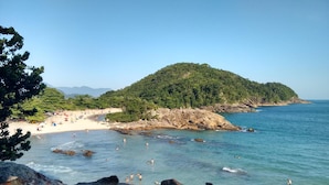A beach with rocky shores, clear blue water, and a forested hill in the background. - Aquarius Chalés (Ubatuba)
