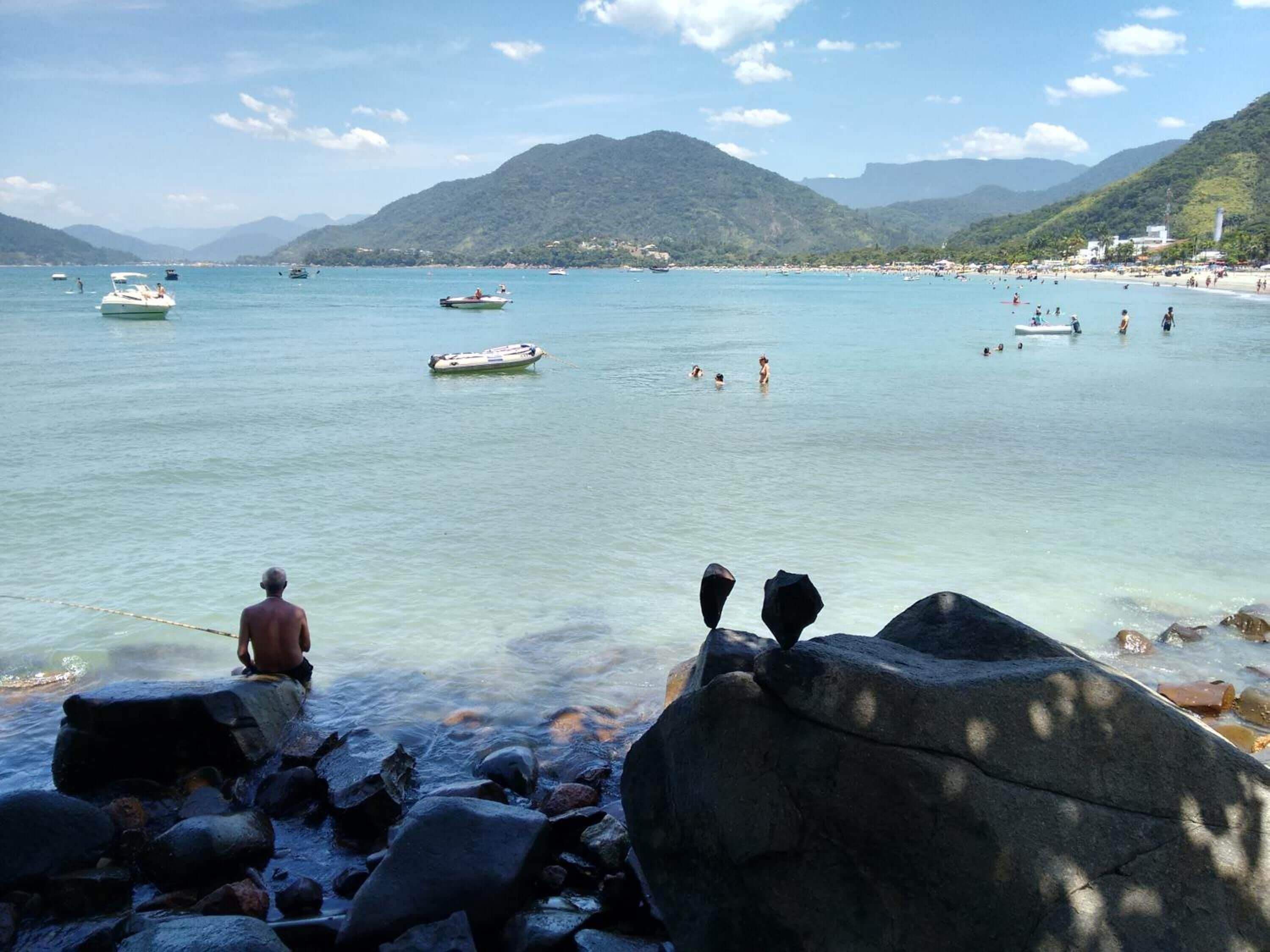 Une plage avec des bateaux, des gens qui se baignent et des montagnes en arrière-plan.