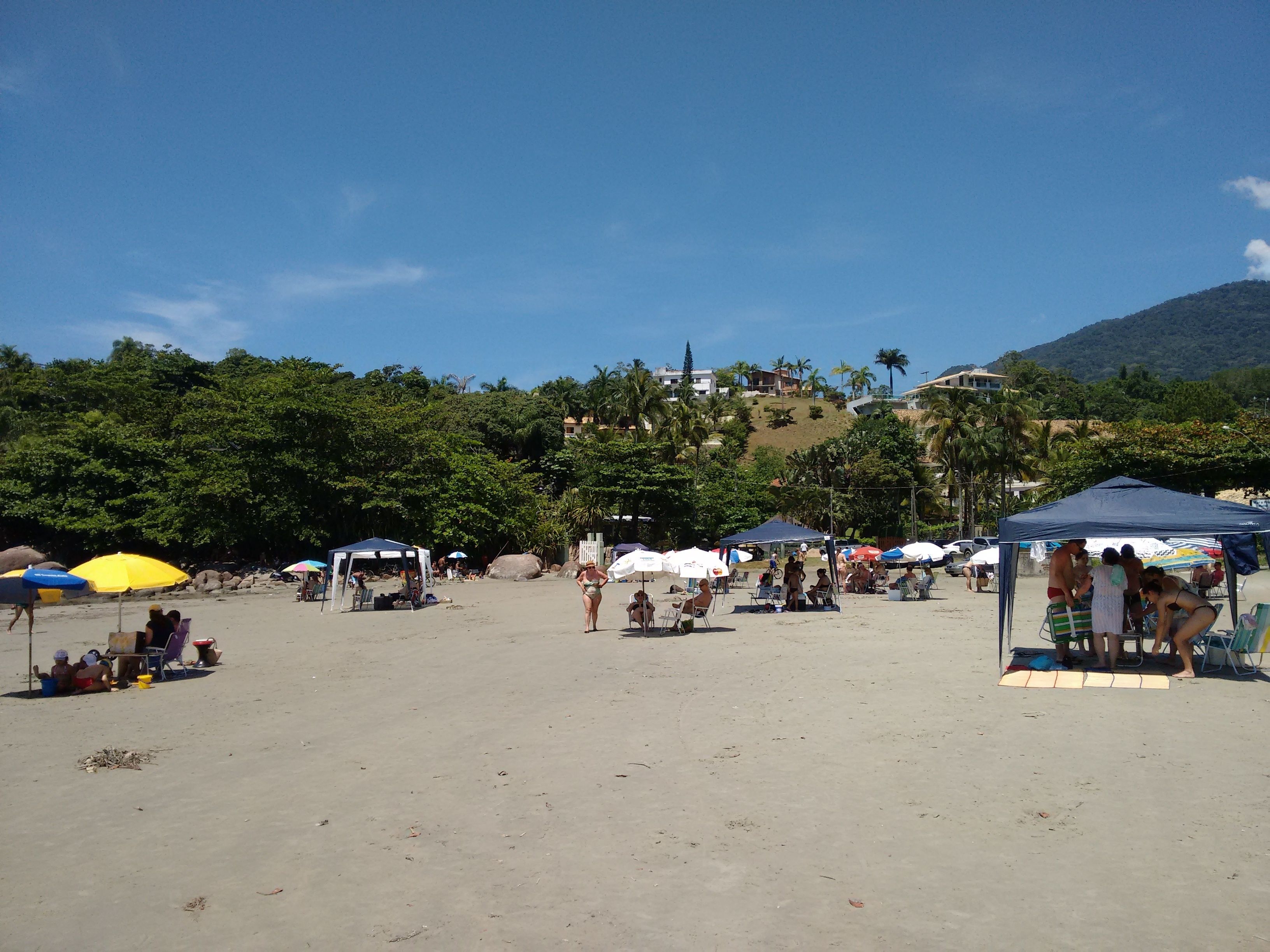 Ein Strand mit Zelten und Sonnenschirmen, Menschen entspannen sich, und ein Hügel mit Gebäuden im Hintergrund.
