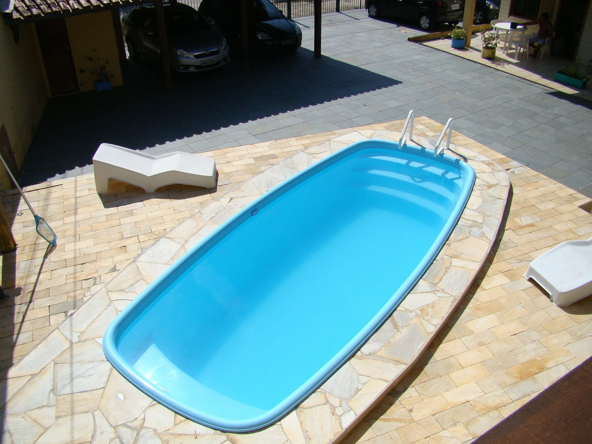 A rectangular swimming pool with a stone border, surrounded by a patio area with chairs and tables.