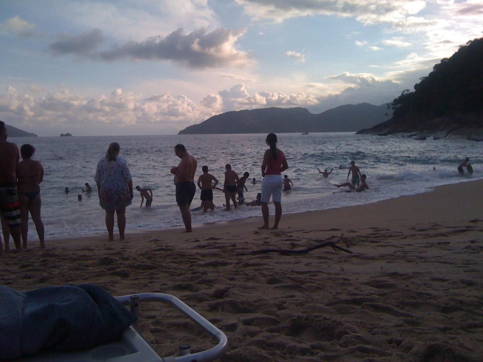 A beach with people swimming and standing on the sand, a boat in the foreground, and a hilly landscape in the background.