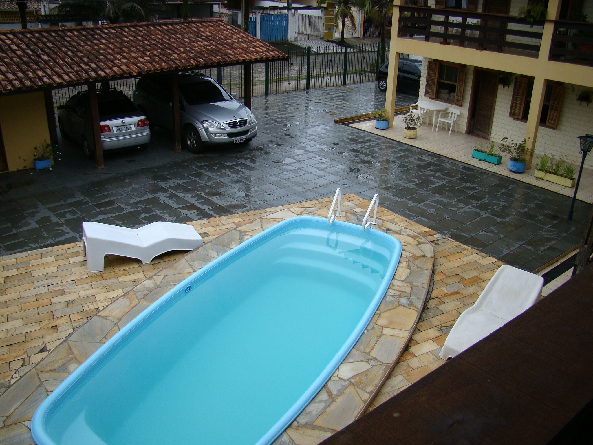 En swimmingpool med fliseterrasse, en overdækket carport og en bygning med altaner.