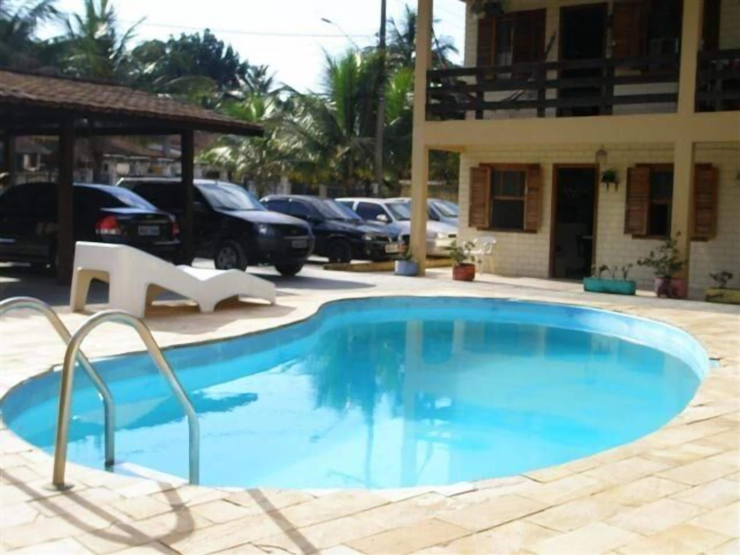 A swimming pool with lounge chairs and a building with multiple cars parked in front.