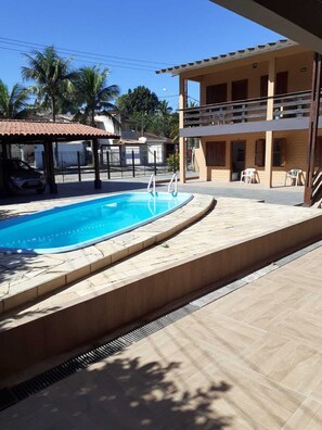 Una piscina con cubierta de piedra, un área de estar techada y un edificio con balcón.