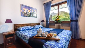 Una cama bien hecha con una colcha estampada de flores, una bandeja con alimentos para el desayuno y una ventana con vista a la montaña.