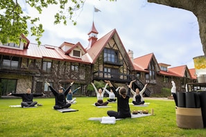 Eine Gruppe von Menschen praktiziert Yoga im Freien auf einer Wiese, im Hintergrund ist ein Gebäude zu sehen.