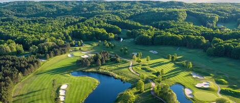 Luchtbeeld van een golfbaan met meerdere bunkers, een vijver en omliggende beboste heuvels.