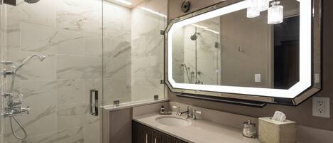 A modern bathroom with a glass-enclosed shower, a large mirror with lights, a sink, and a tissue box.