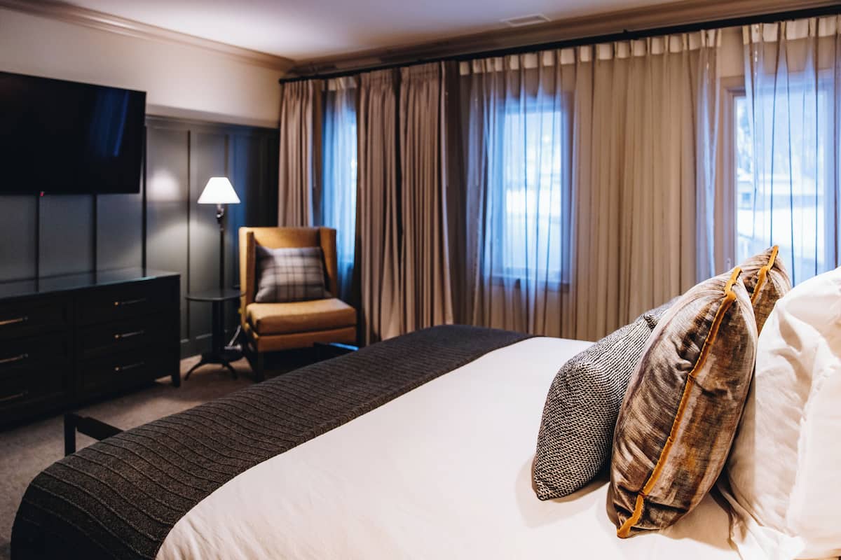 Ein Hotelzimmer mit einem großen Bett, einem an der Wand montierten Fernseher, einem Sessel und Vorhängen.