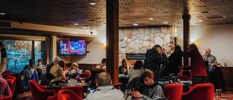 A restaurant with red chairs, tables, a stone wall, and a television screen.