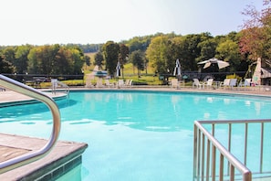 Ein Pool mit Metallgeländer, umgeben von weißen Liegestühlen und Sonnenschirmen, mit Bäumen und einem Gebäude im Hintergrund.