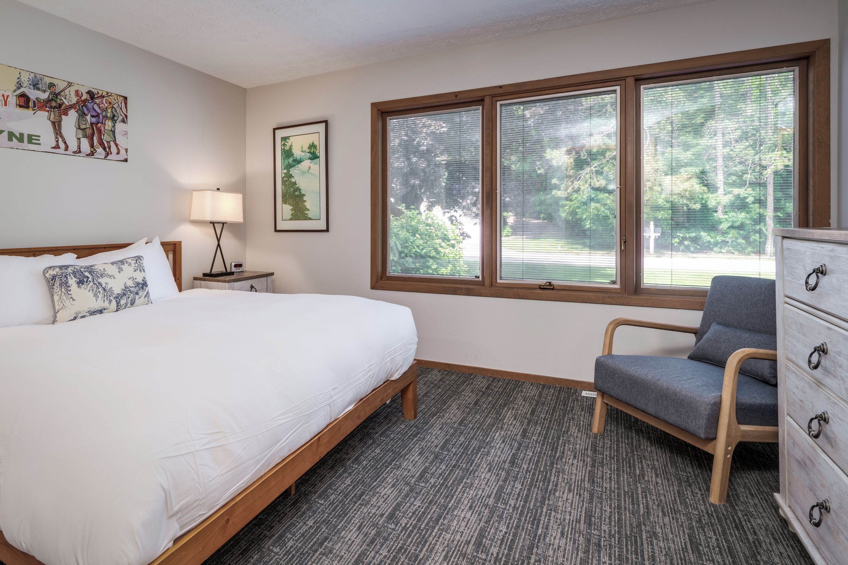 Une chambre à coucher comprenant un lit, une chaise, une commode, une lampe et une fenêtre donnant sur des arbres.