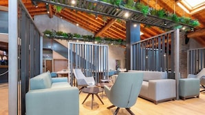 Un área de descanso moderna con sofás y sillones, piso de madera, y un techo con vigas a la vista y plantas.