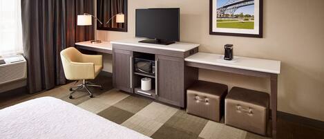 A hotel room with a bed, a desk with a television, a chair, a lamp, a picture frame, and storage boxes.