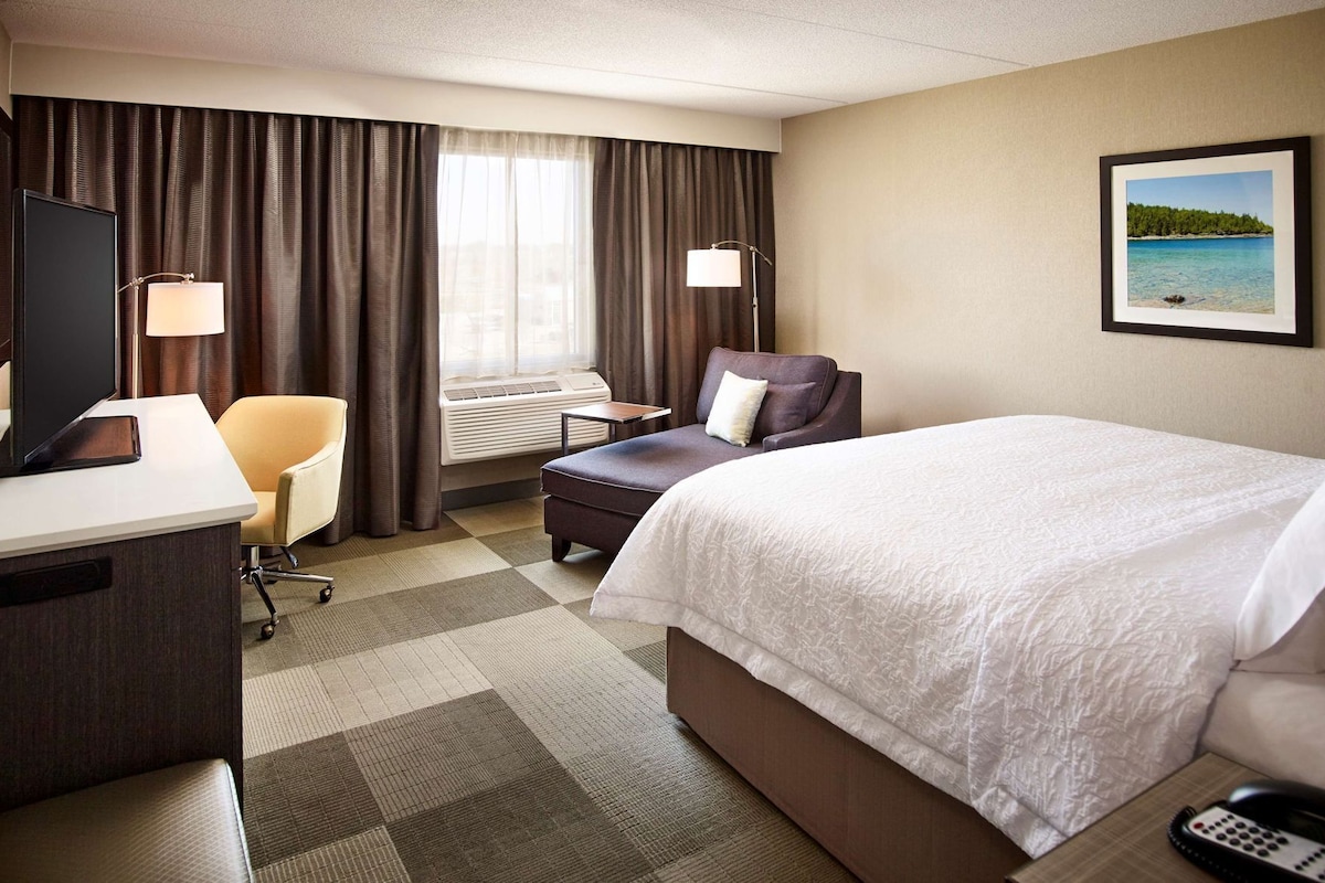 A hotel room with a bed, desk, chair, and a window with curtains.