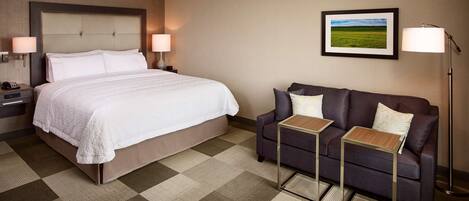 Habitación de hotel con cama, sofá, dos mesitas auxiliares, lámpara, un cuadro enmarcado y un teléfono en la mesita de noche.
