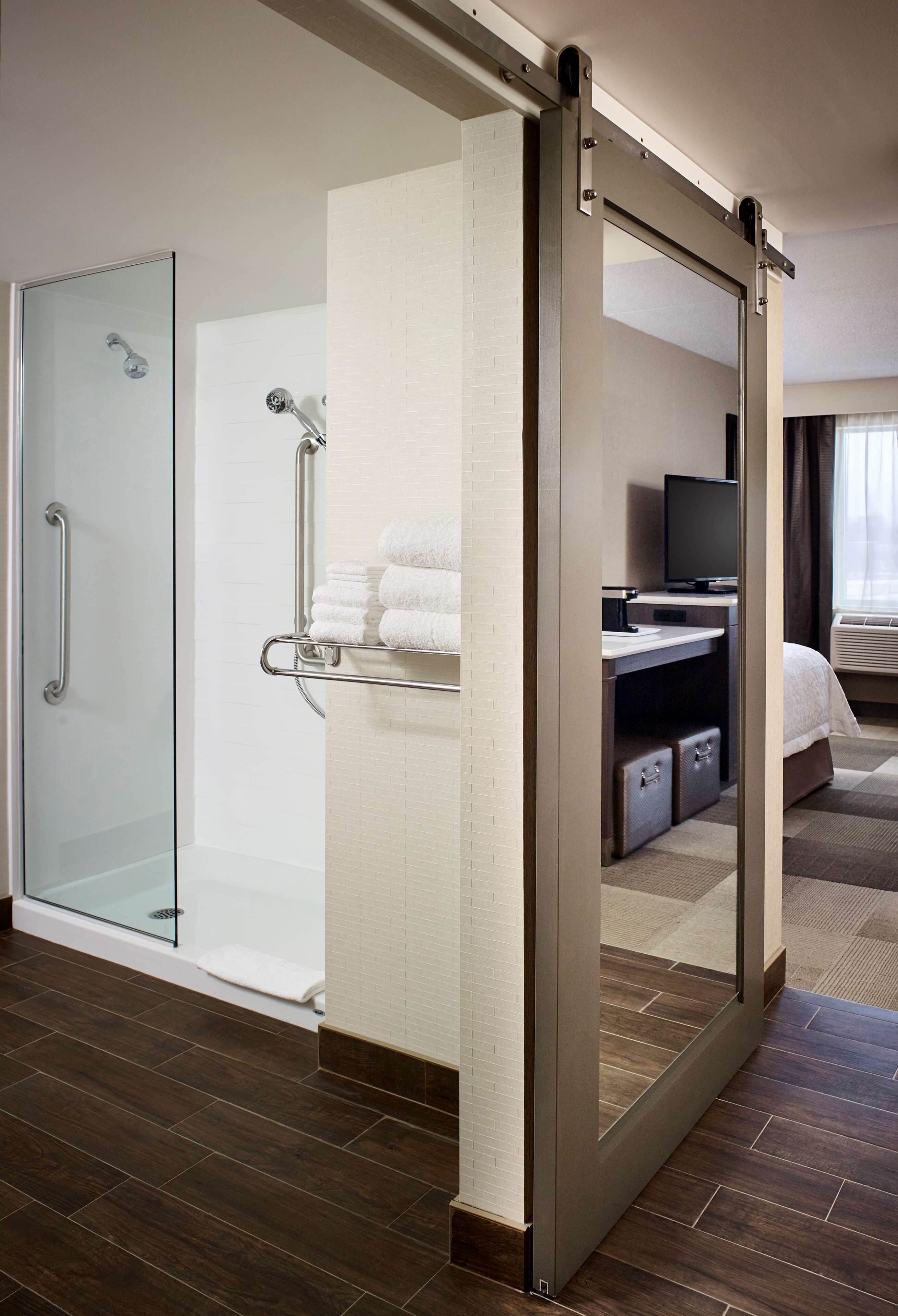 A hotel room with a glass-enclosed shower, a bed with a TV, and a large mirror.