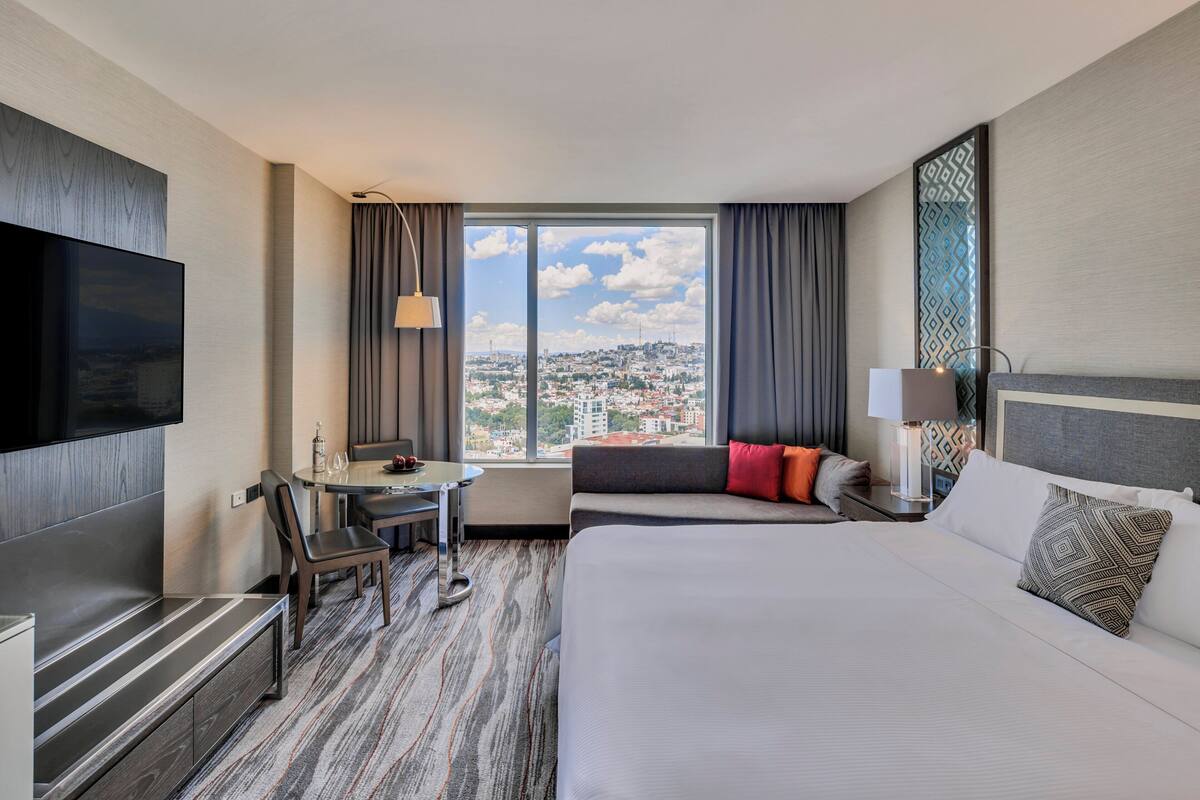 A hotel room with a large bed, a desk with a chair, a TV, and a window with a city view.