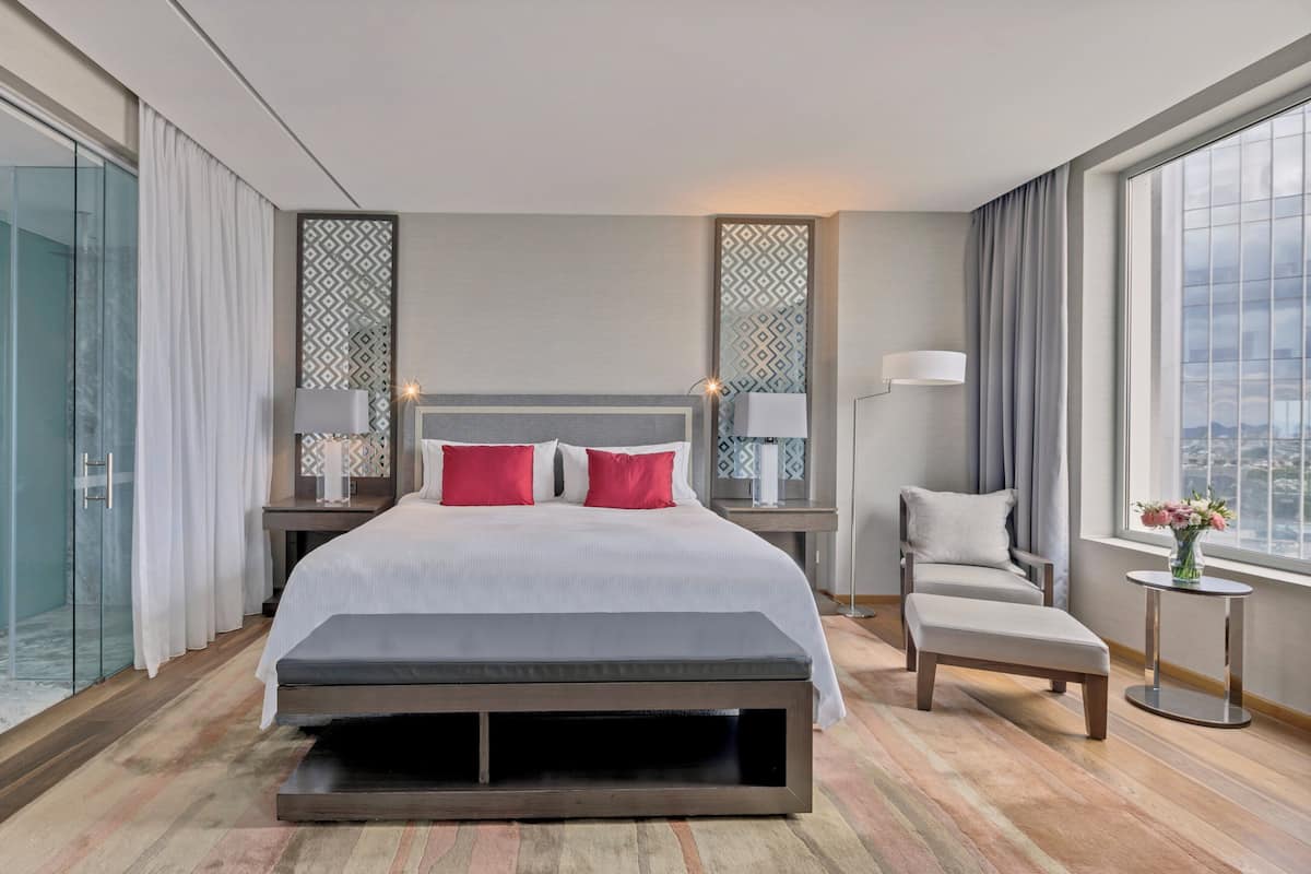 A modern hotel room with a large bed, a bench, a chair, a bedside table with lamps, and a view of the city through large windows.