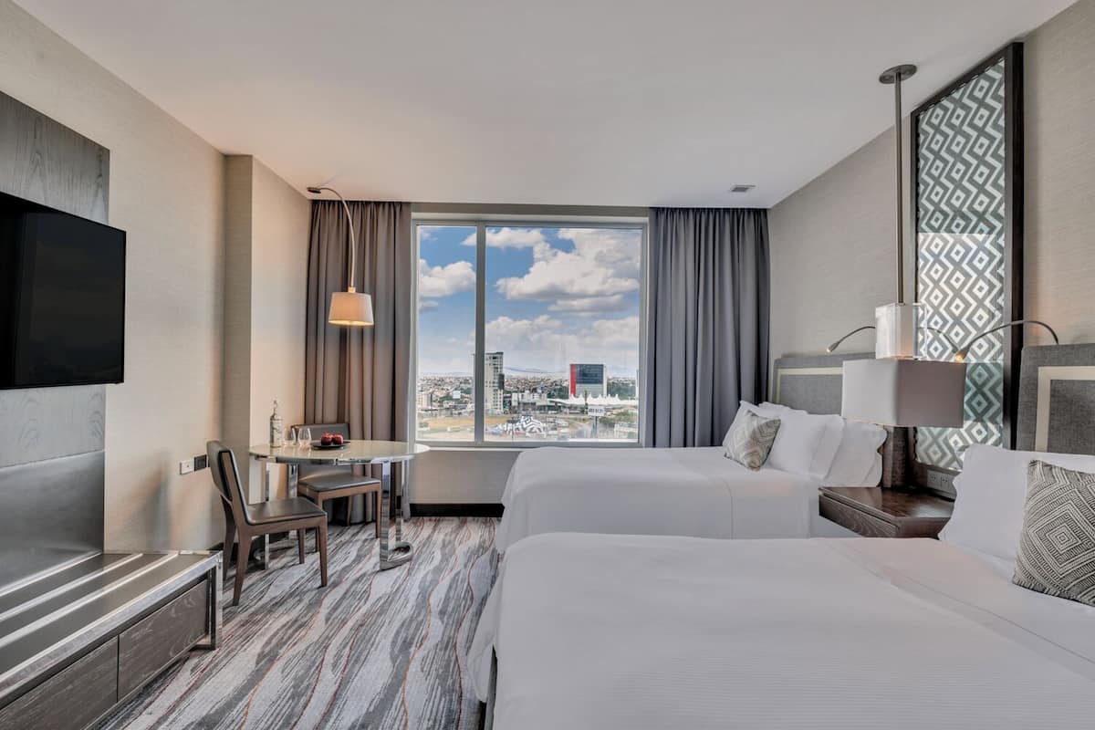 A hotel room with a bed, a desk with a chair, a TV, and a large window with a city view.