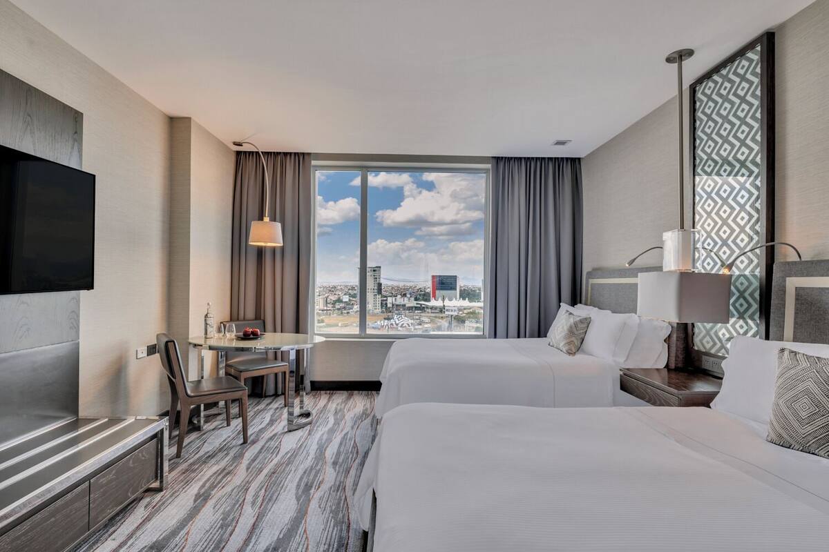 Habitación de hotel con cama, escritorio con silla, televisión y una ventana grande con vista a la ciudad.