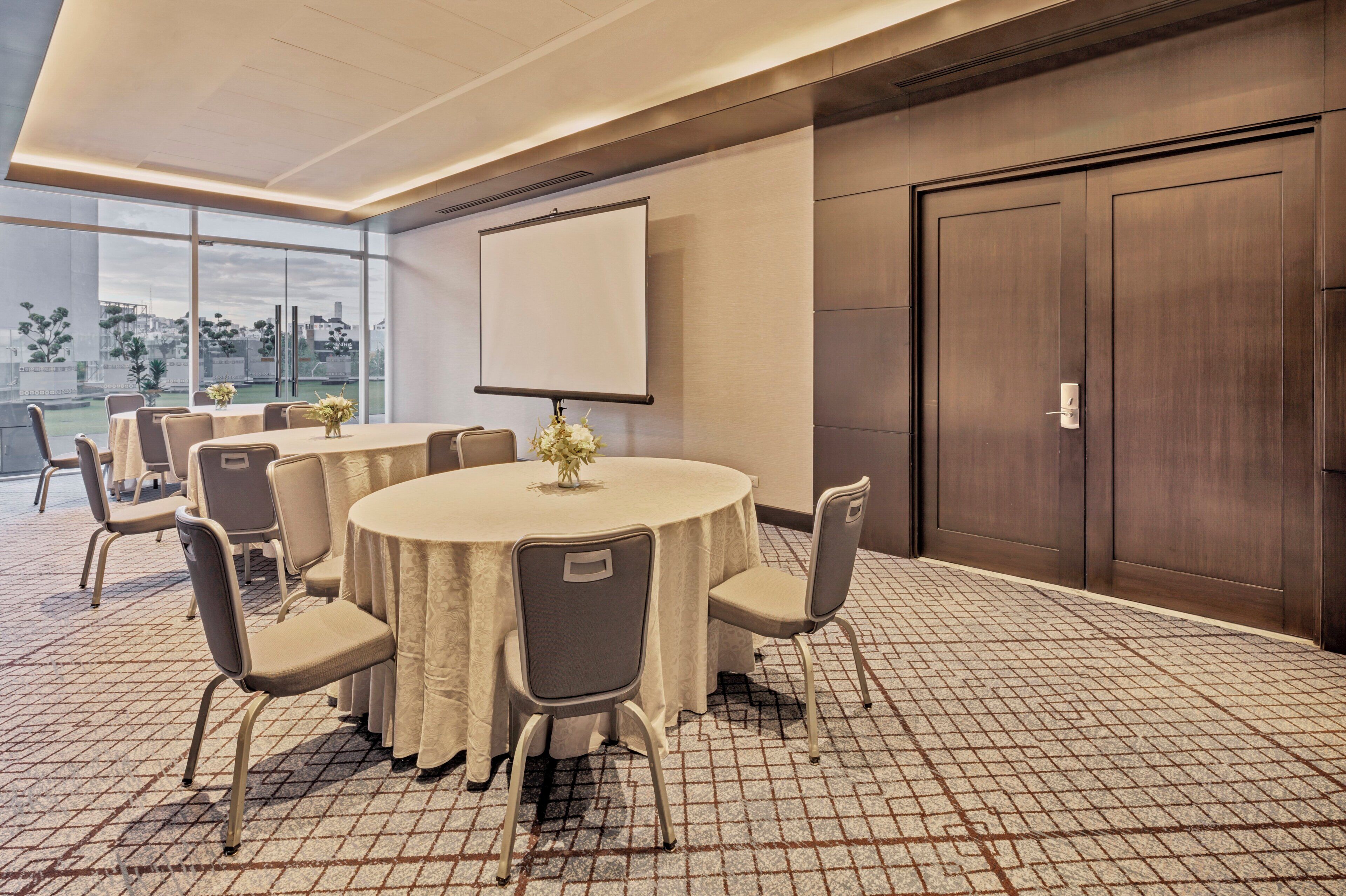 Un salón de conferencias con una gran mesa, sillas, una pantalla de proyector y vistas al exterior.