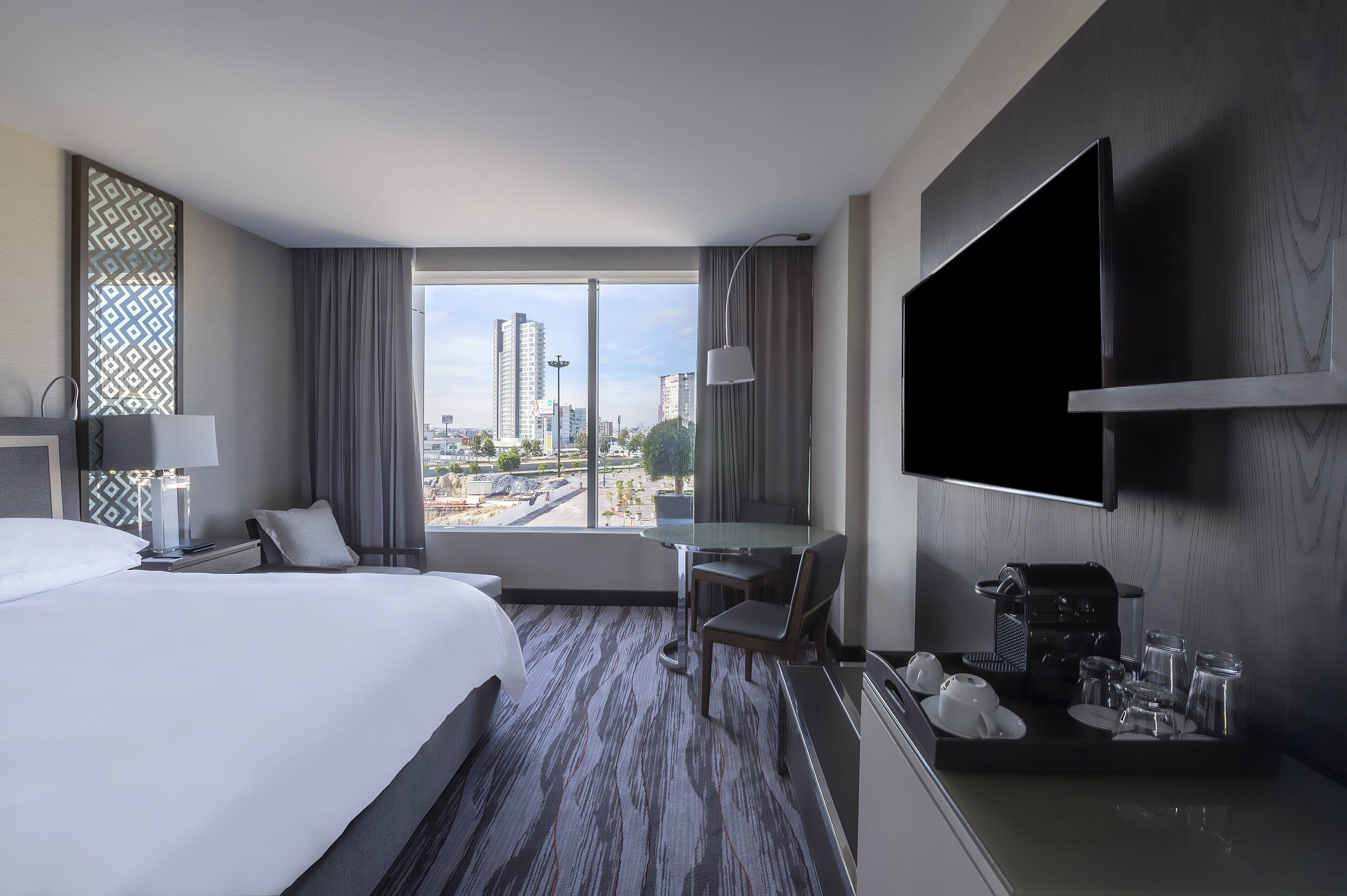 Habitación de hotel con una cama grande, un escritorio, una silla, un televisor y vistas a la ciudad.