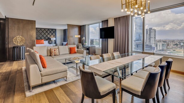 Een moderne hotelkamer met een grote eettafel, comfortabele zithoek, een flatscreen televisie en uitzicht over de stad door grote ramen.