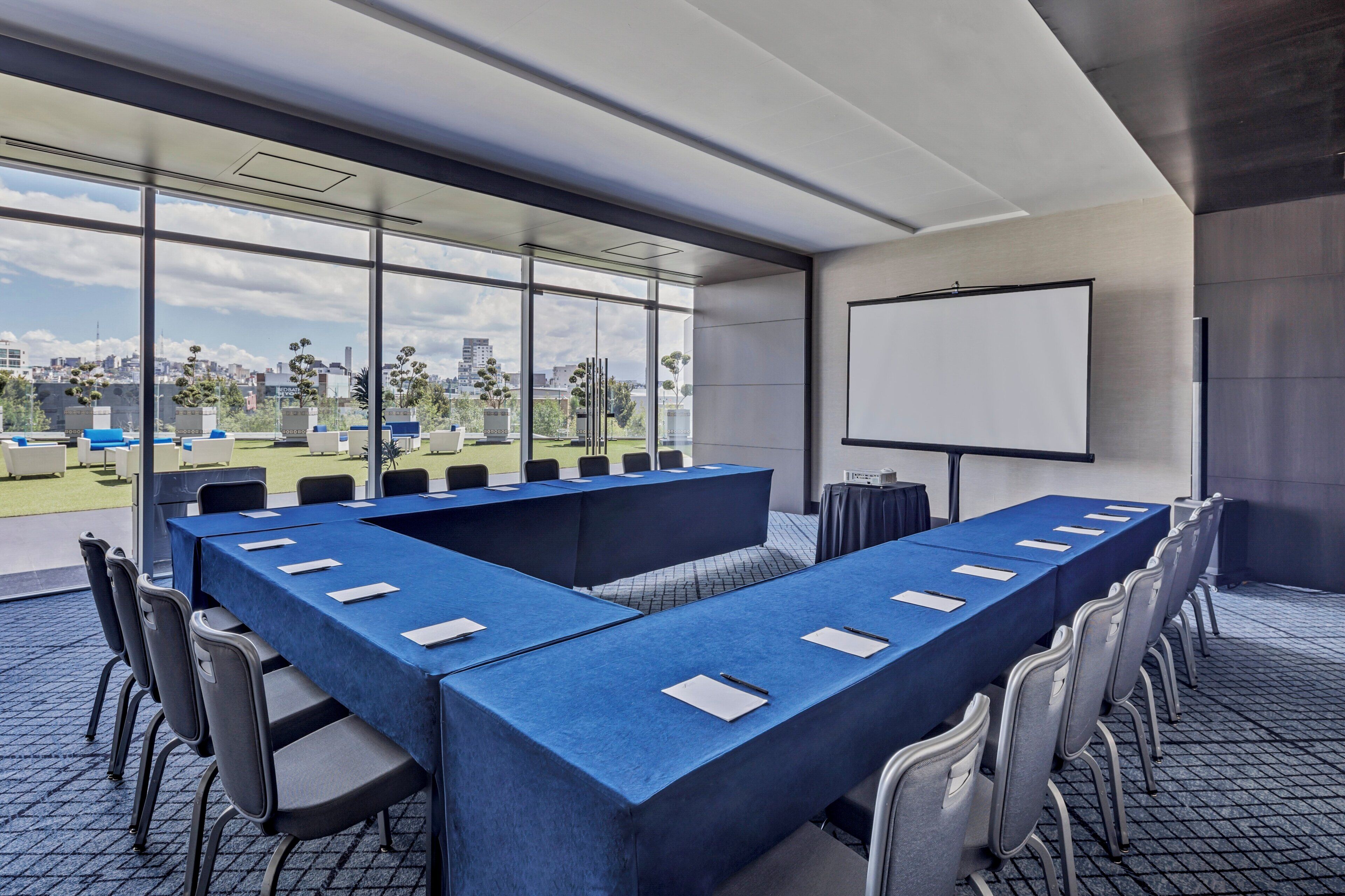 Una sala de conferencias moderna con una mesa larga, sillas, una pantalla de proyector y ventanas amplias con vistas a la ciudad.