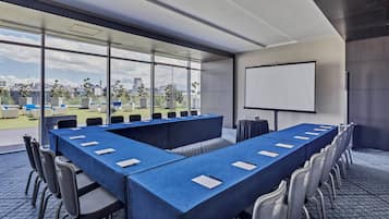 Una sala de conferencias moderna con una mesa larga, sillas, una pantalla de proyector y ventanas amplias con vistas a la ciudad.