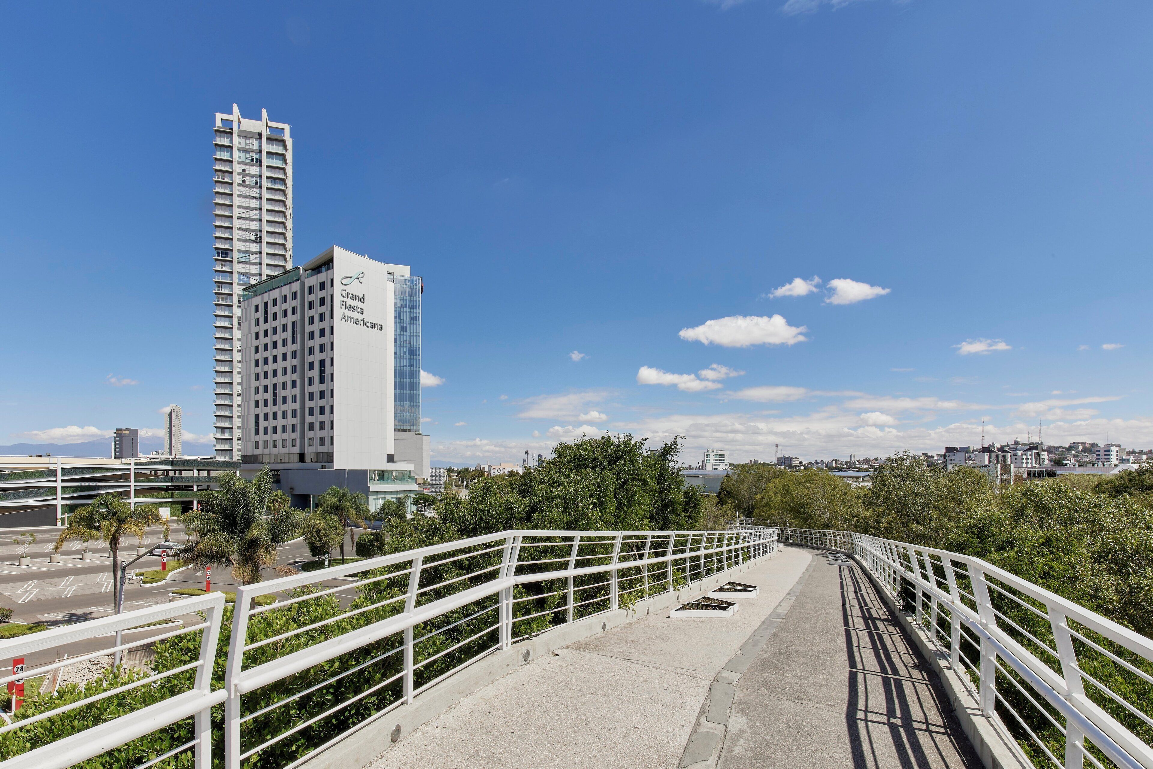 Uma ponte para pedestres com corrimão branco, levando a um edifício moderno com fachada de vidro e a placa 'Grand Fiesta Americana'.