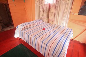 A single bed with a striped bedspread and pillows, a green rug, and a window with floral curtains. - Beach Road Cottages at Jah Bs (Negril)
