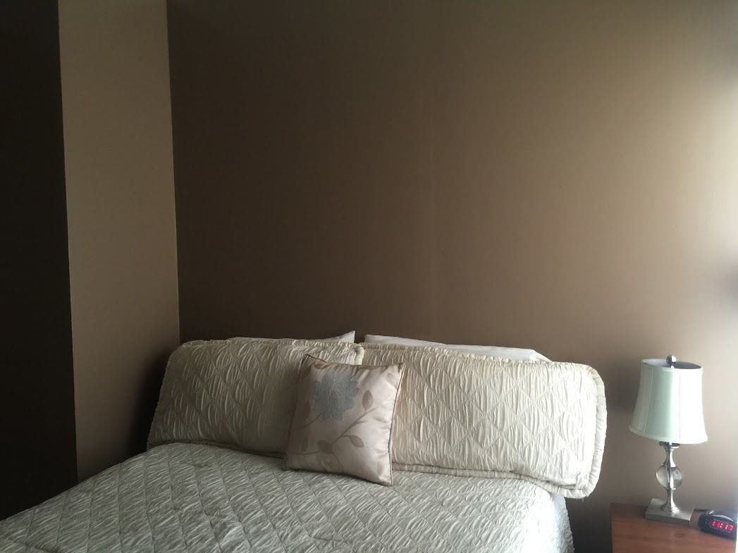 Una cama con cubrecama acolchado y almohada estampada, una lámpara de noche y un reloj sobre una mesita de madera.