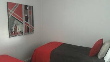 Ein Zimmer mit zwei Einzelbetten, jeweils mit einer roten und grauen Decke sowie einem roten Kissen. An der Wand hängt ein gerahmtes Bild.