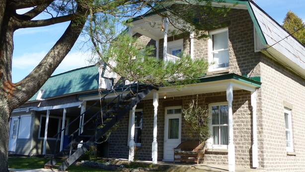 A two-story stone building with a green roof, a front porch, and a staircase leading to the second floor. - Hôtel du Chasseur (Brownsburg-Chatham)