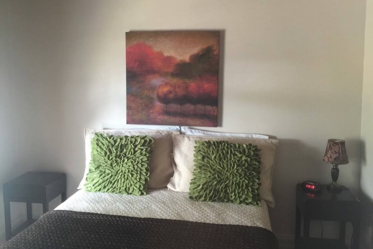 Un dormitorio con una cama, dos almohadas verdes, una mesita de noche con una lámpara y un cuadro en la pared.