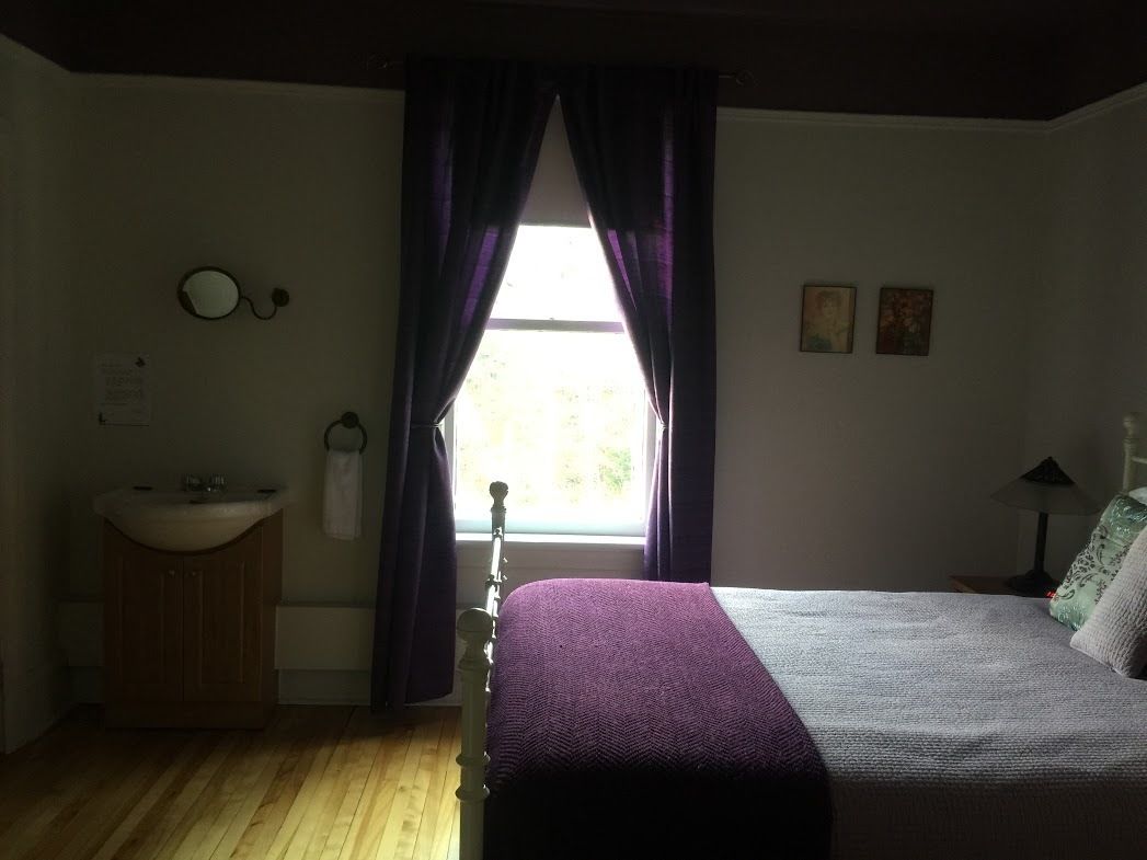 A bedroom with a bed, a window with curtains, a bedside table, a lamp, and a wall-mounted mirror.