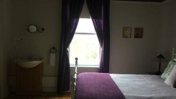 A bedroom with a bed, a window with curtains, a bedside table, a lamp, and a wall-mounted mirror.