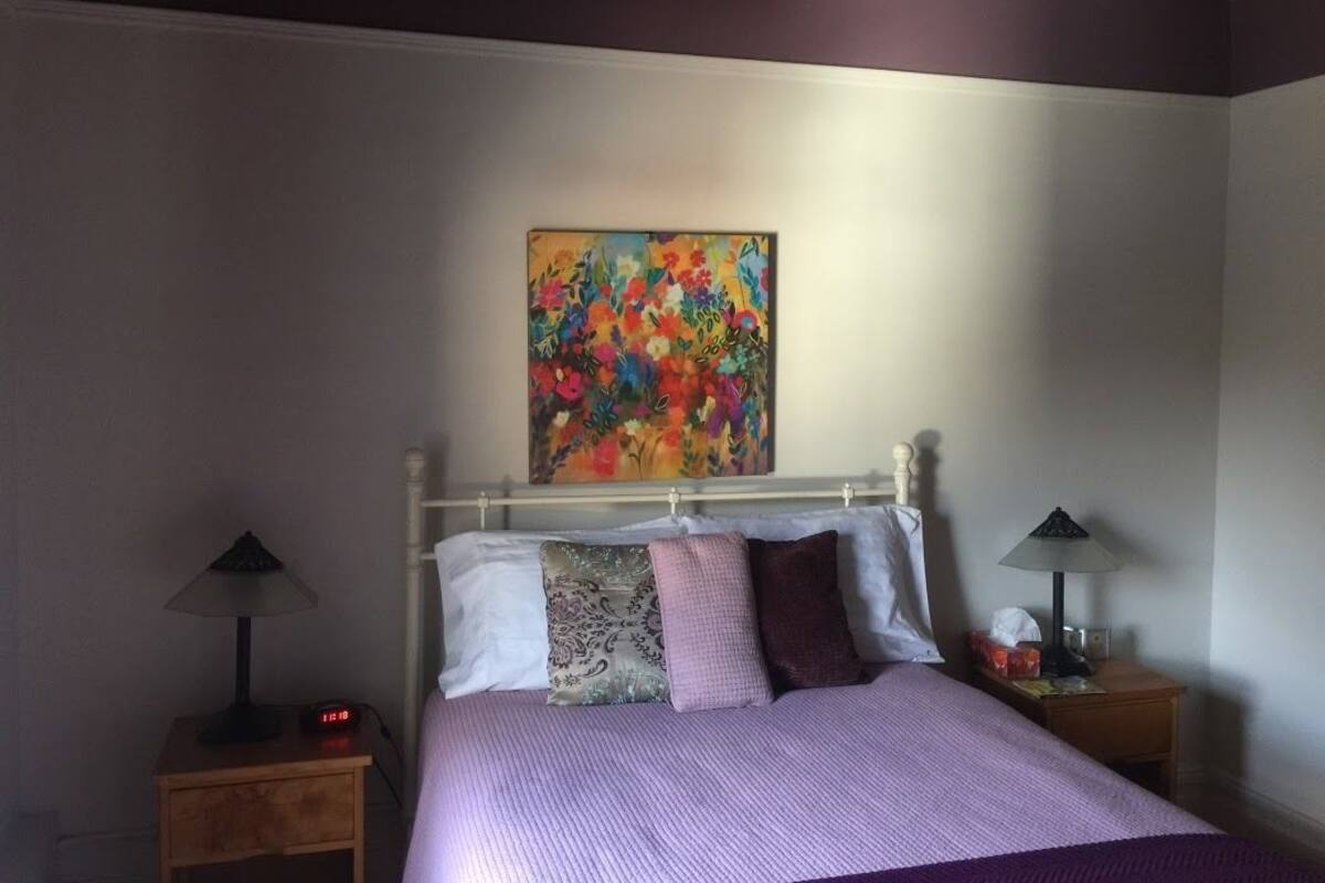 Un dormitorio con una cama, dos mesitas de noche, una lámpara en cada una y un cuadro llamativo en la pared.