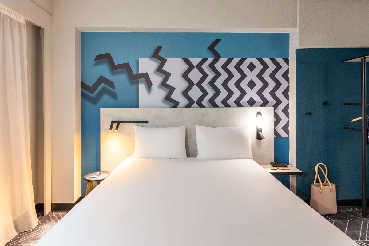 A hotel room with a large bed, a headboard with a geometric wall design, a bedside table with a lamp, and a door with a handle.