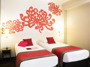A hotel room with two beds, each with a red pillow and a red blanket. There is a nightstand with a lamp and a phone on it. The walls have decorative red patterns. - Ibis Styles Bordeaux Meriadeck (Bordeaux)
