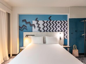 Una habitación de hotel con una cama grande, un cabecero con diseño geométrico en la pared, una mesita de noche con lámpara y una puerta con pomo.