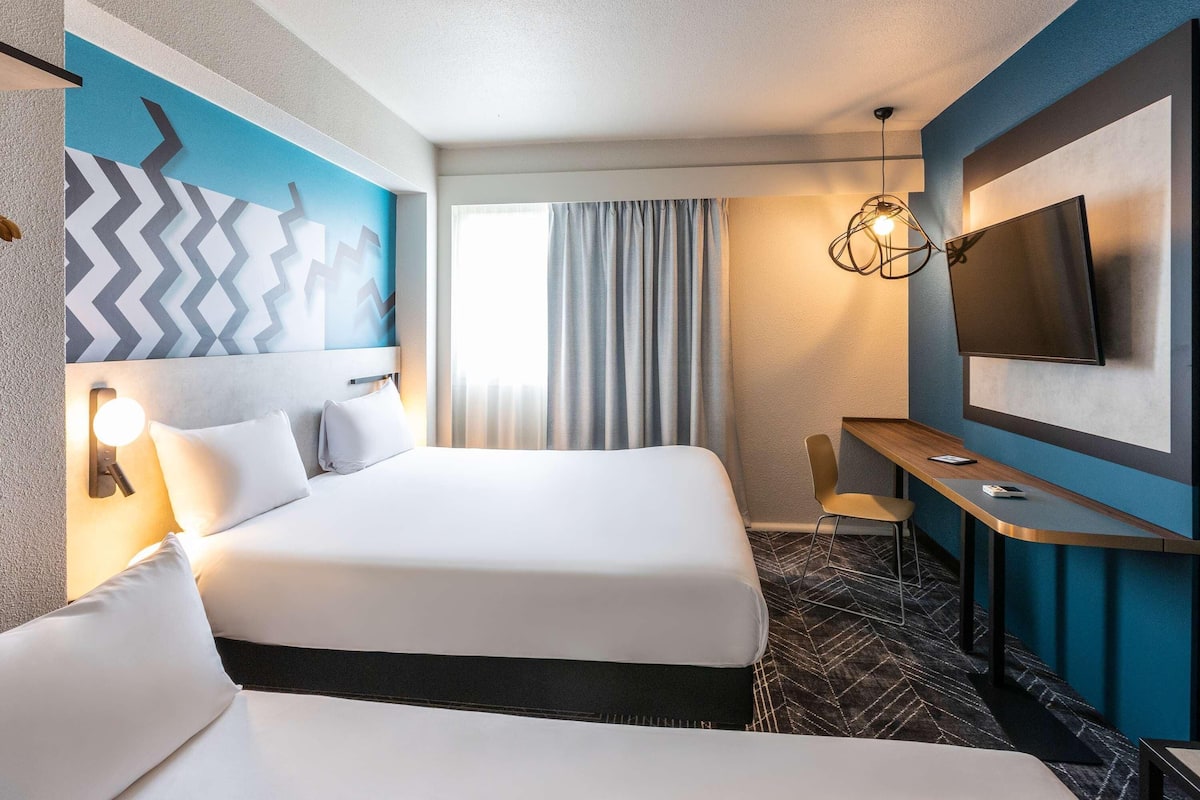 A hotel room with a large bed, a desk, a TV, and a modern light fixture.