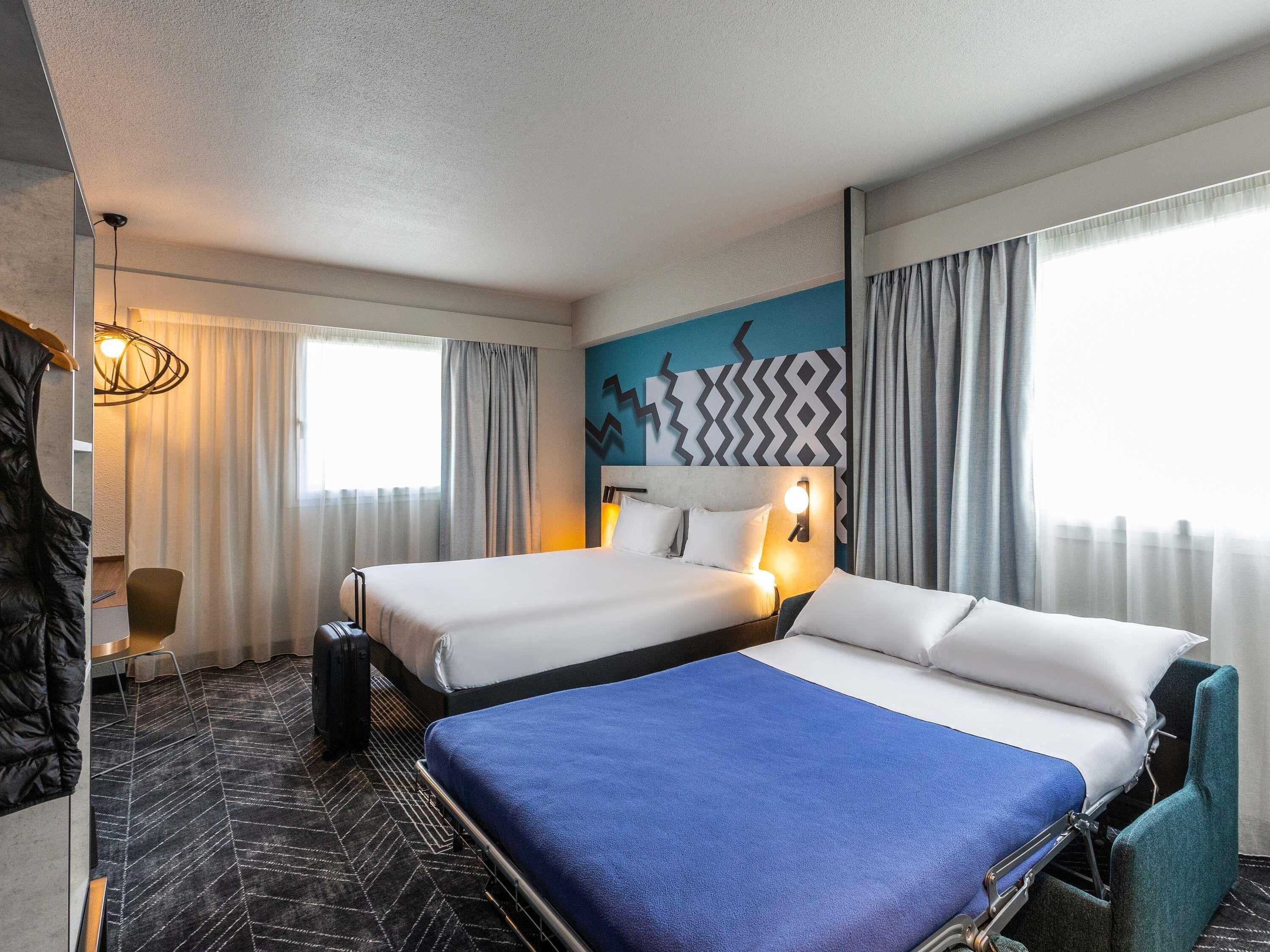 Habitación de hotel con dos camas, un escritorio, una silla y un amplio ventanal con cortinas.