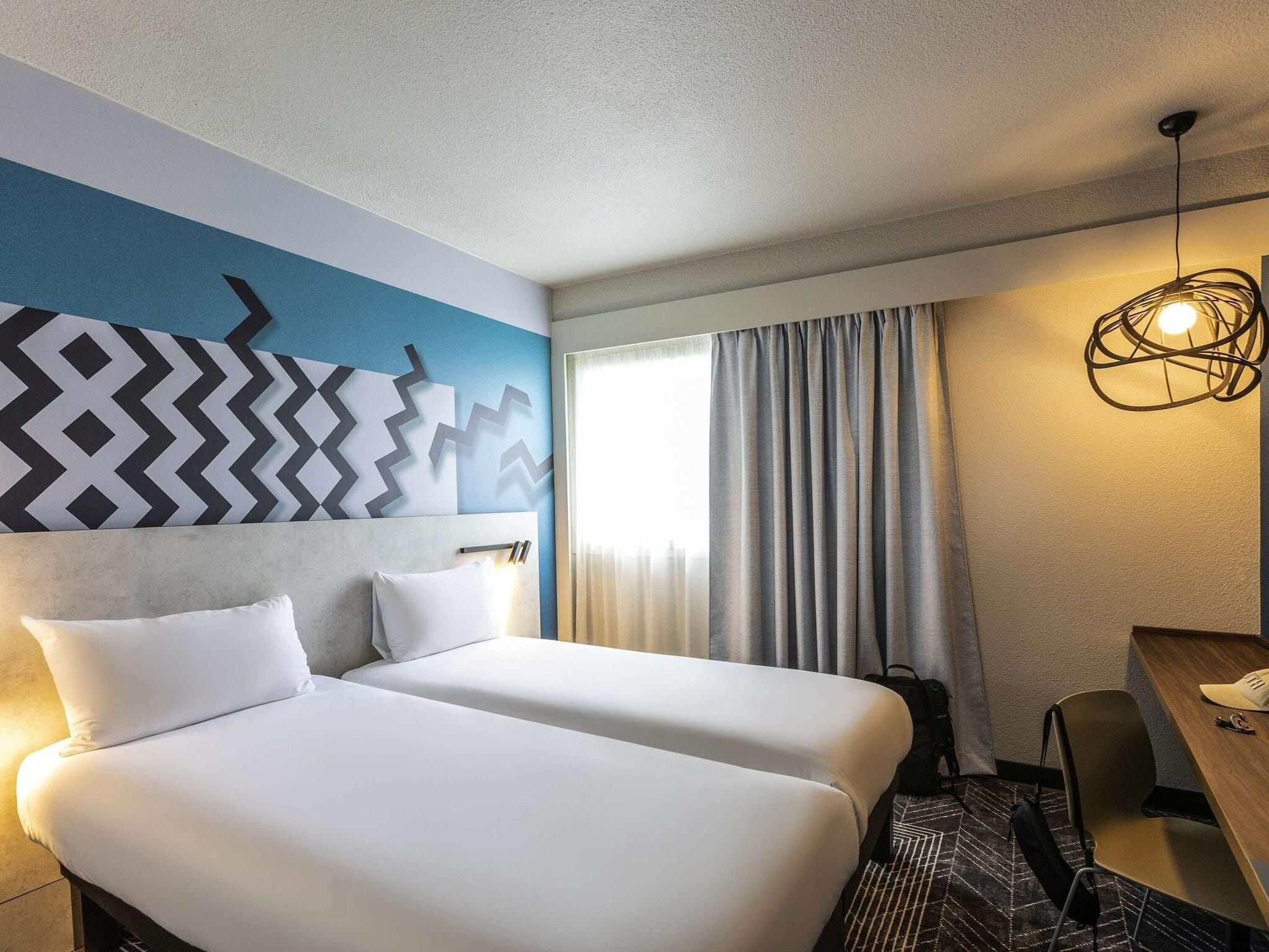 Habitación de hotel con dos camas, un escritorio, una silla, una lámpara y una ventana con cortinas.