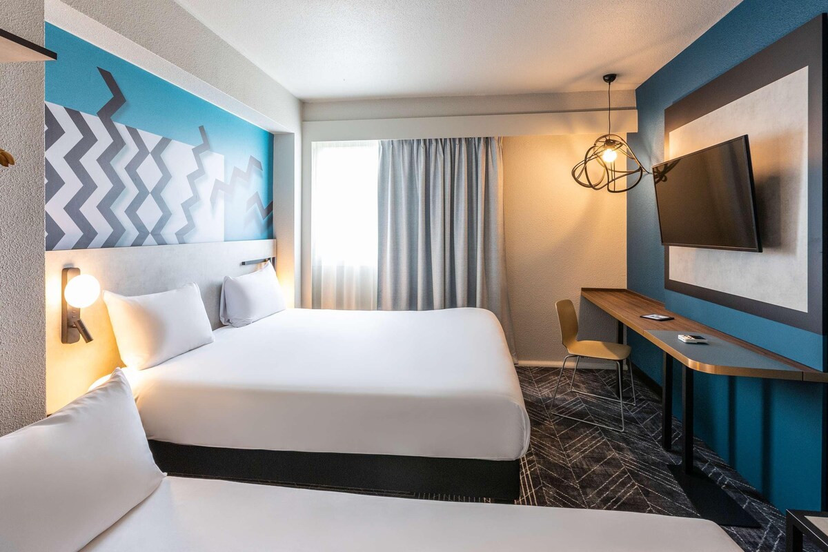 A hotel room with a large bed, a desk, a TV, and a modern light fixture.