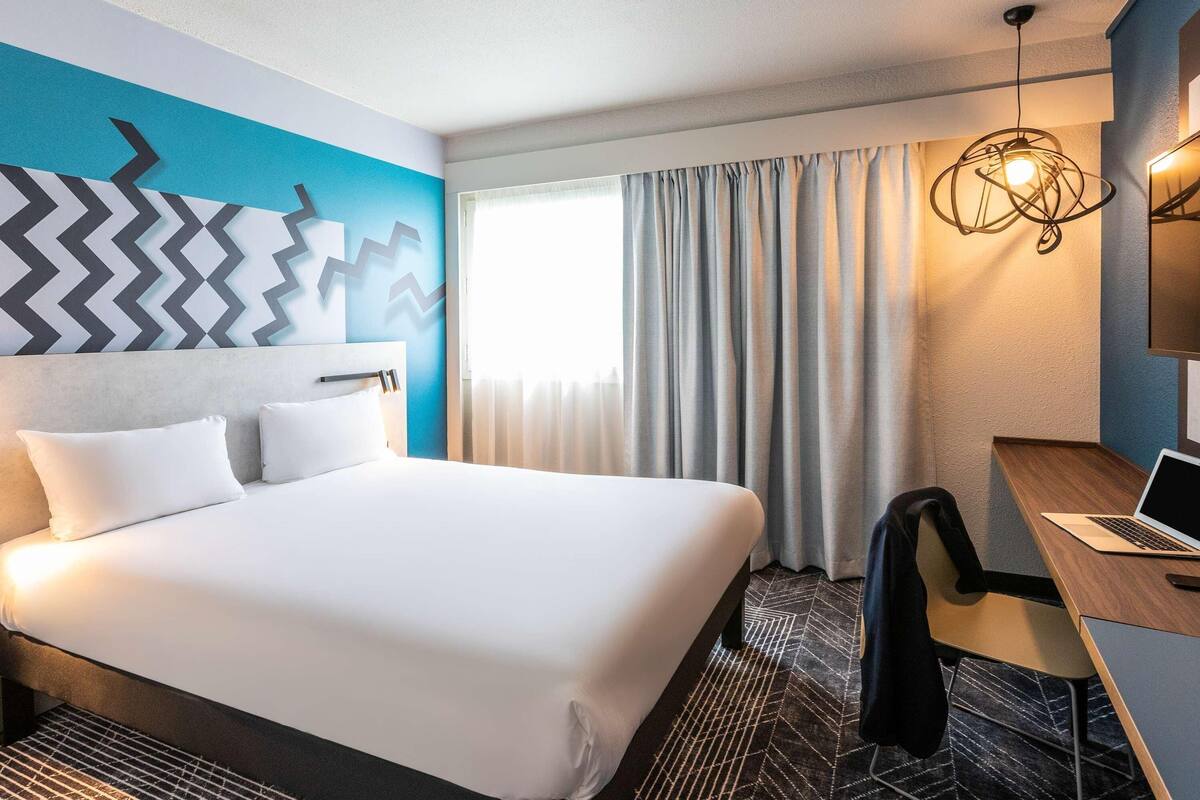A hotel room with a large bed, a desk with a laptop, a chair, and a modern light fixture.