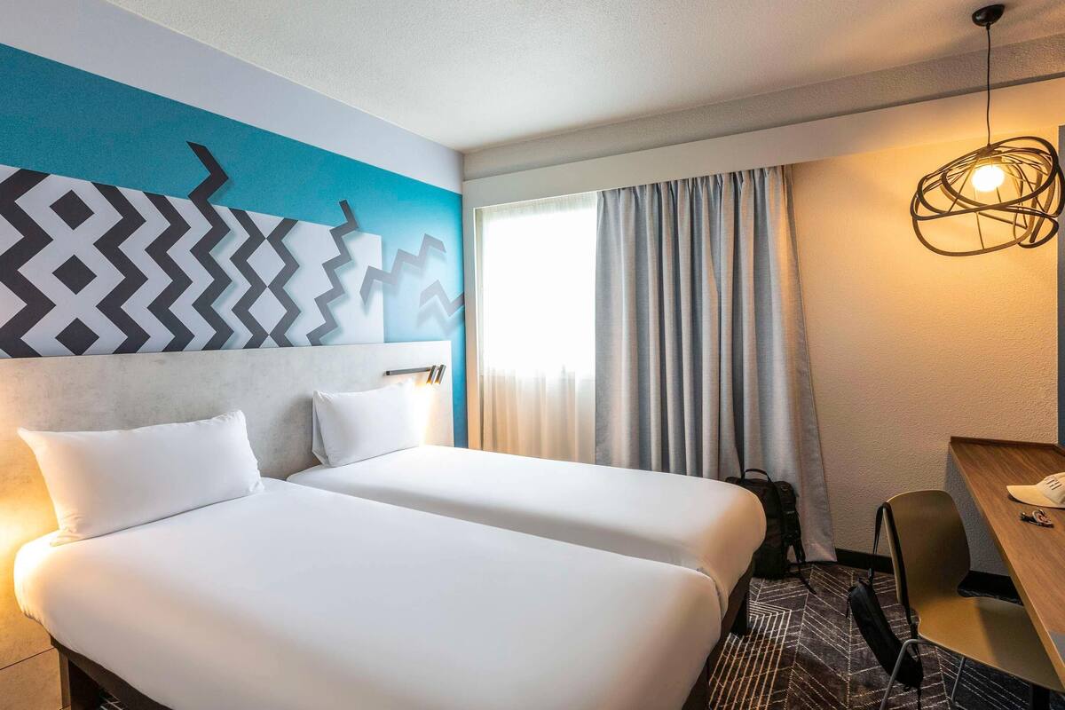 A hotel room with two beds, a desk, a chair, a patterned rug, and a modern light fixture.