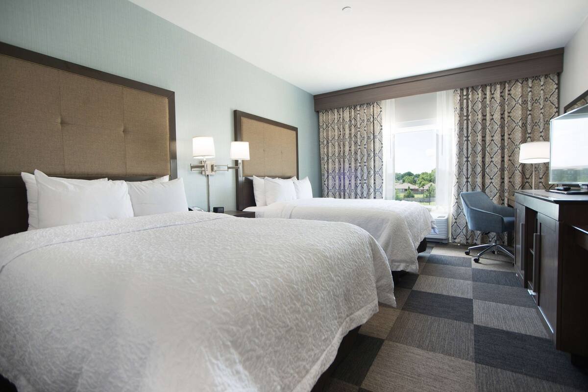 A hotel room with two beds, a desk, a chair, and a large window with curtains.