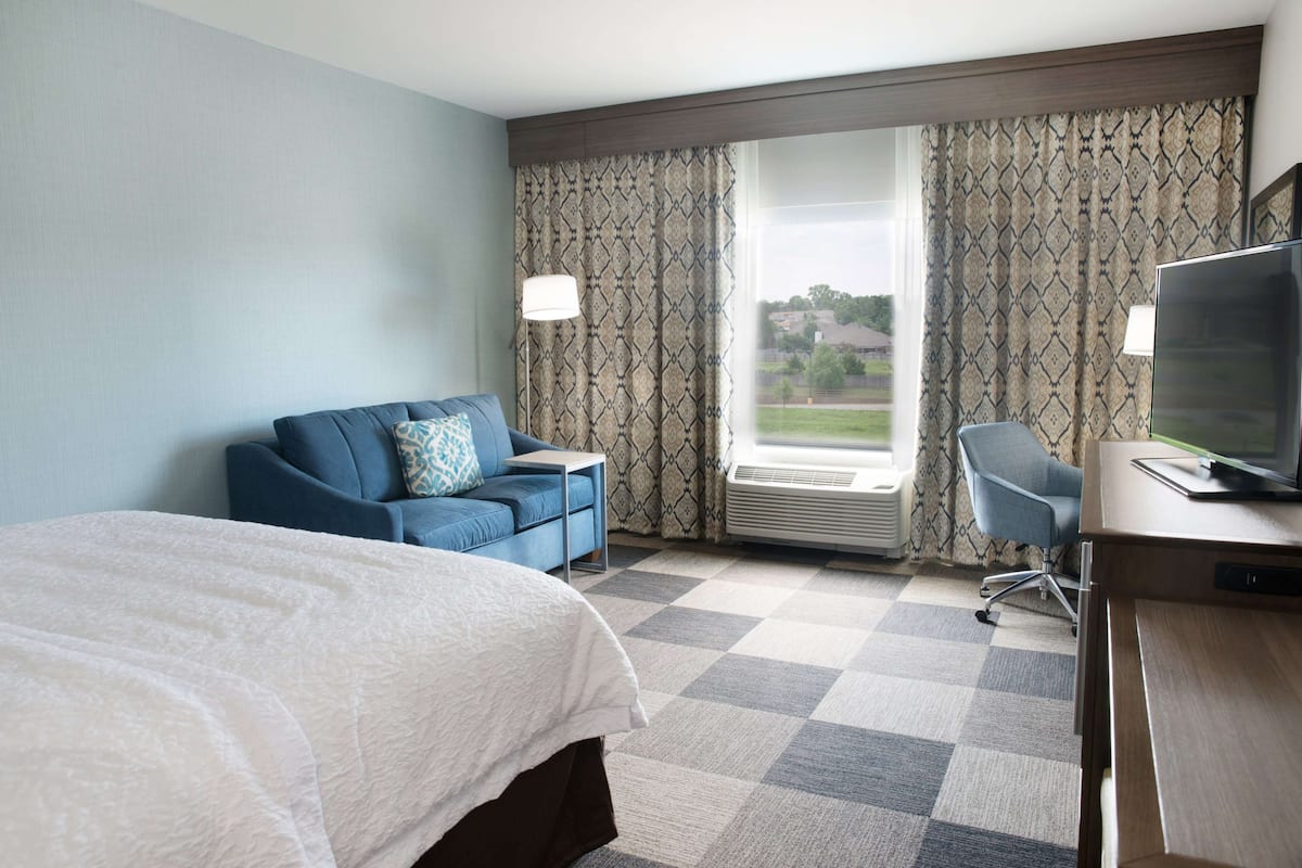 A hotel room with a bed, a blue sofa, a desk with a computer, and a large window with patterned curtains.